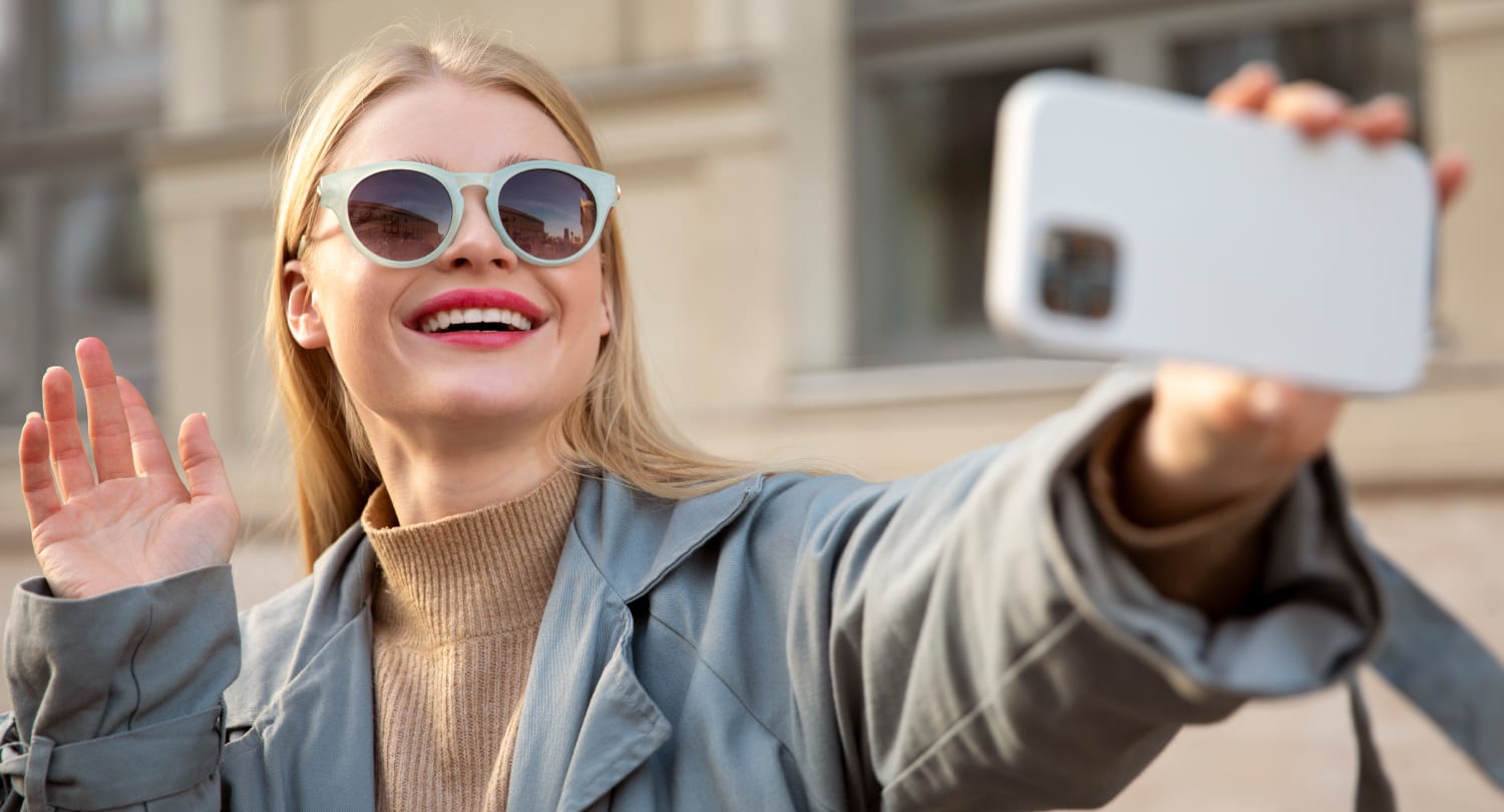 Smiling woman wearing stylish sunglasses taking a selfie with a smartphone, showcasing eyewear fashion and social media engagement.